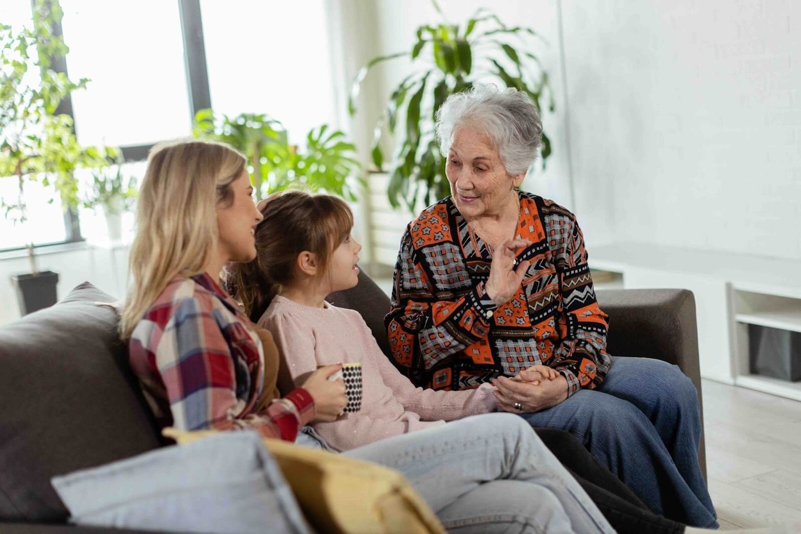  Three generations of family members—a grandmother, her daughter, and granddaughter—sit together on a couch sharing stories and laughter, capturing meaningful family history moments and creating lasting memories across generations.