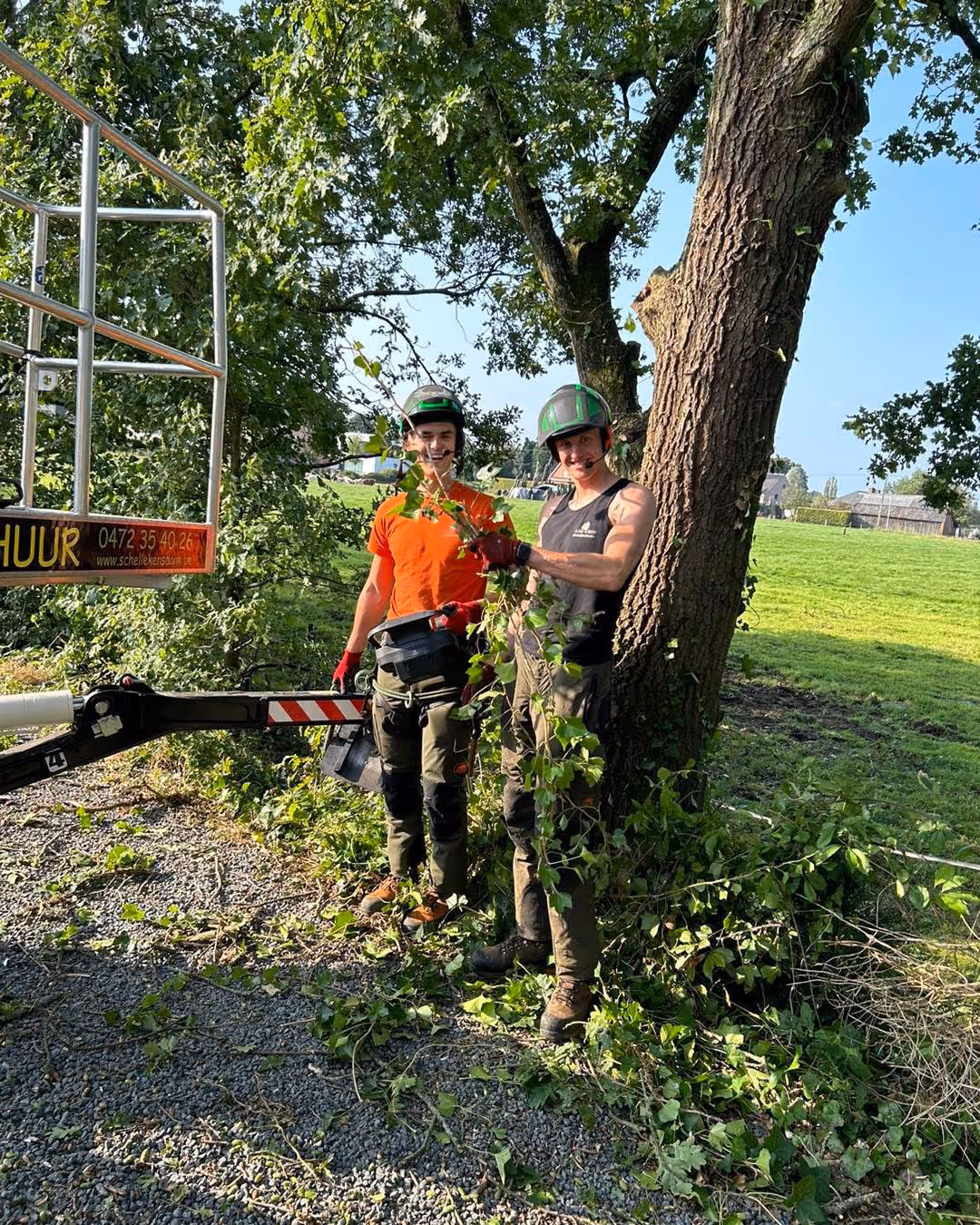 Twee boomverzorgers in beschermende kleding glimlachen terwijl ze takken van een grote boom snoeien. Ze staan op een grindpad met een groen veld op de achtergrond.