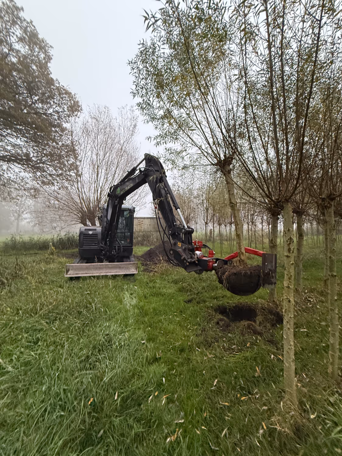 Een graafmachine in een mistig veld ontwortelt een boom met zijn schep, waardoor klonten aarde zichtbaar worden. De omringende bomen en mist creëren een serene, natuurlijke sfeer.