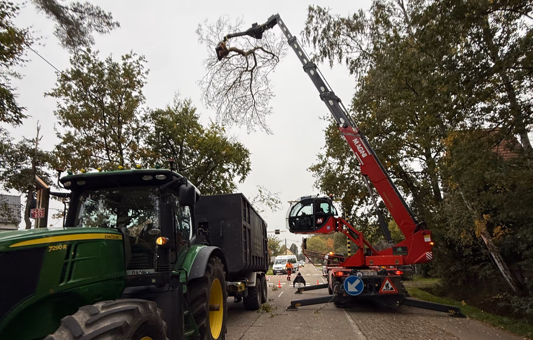 Een straat met een groene tractor en een aanhangwagen geparkeerd naast een rode kraan die een boomtak optilt. Werknemers in veiligheidskleding regelen het verkeer, terwijl bomen het tafereel omringen.