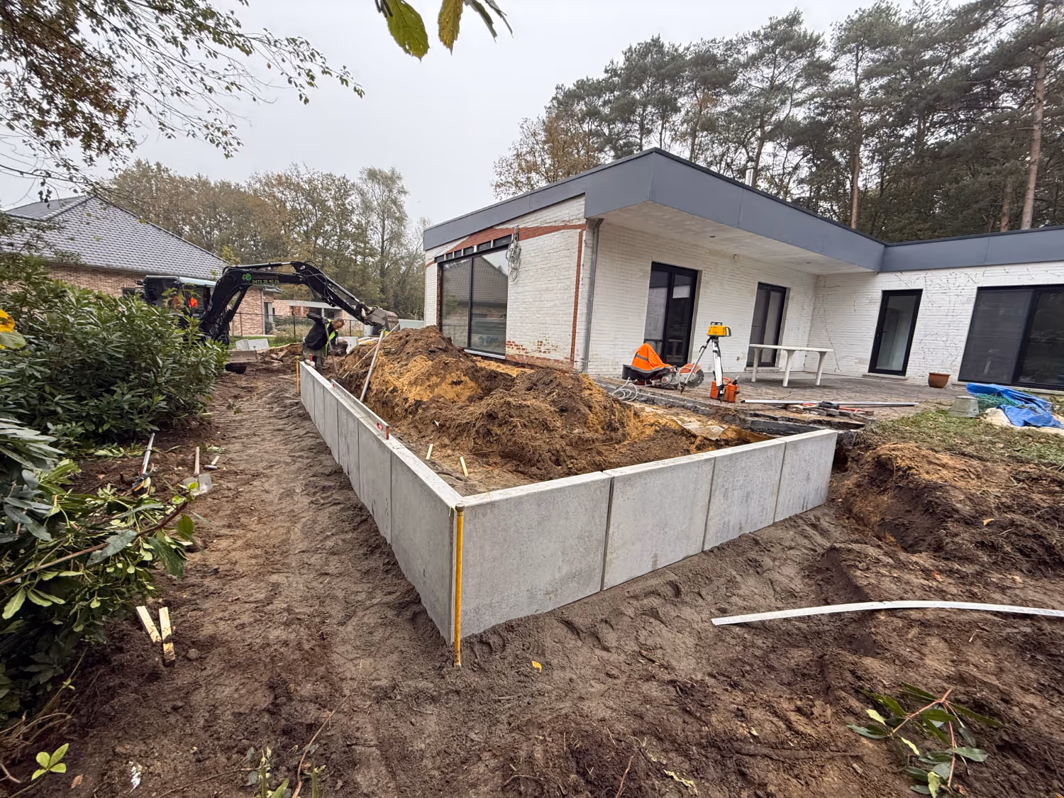 Op een bouwplaats naast een modern wit huis is een uitgegraven gebied met metalen panelen te zien. Er zijn arbeiders en machines zichtbaar, omringd door bomen.