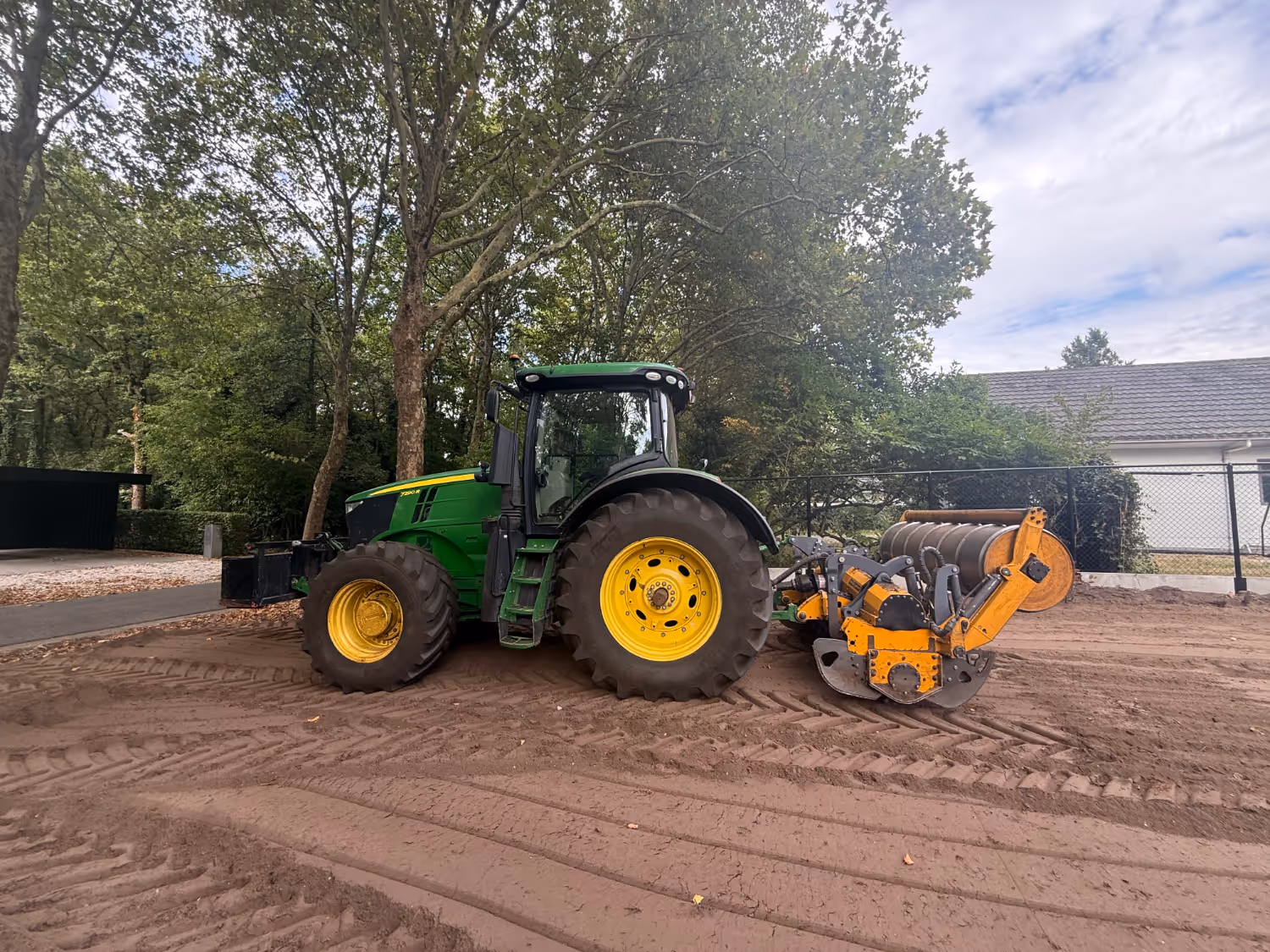 Een groene tractor met gele accenten staat op een onverhard pad, met een grote wals als aanbouwdeel. Op de achtergrond staan bomen en een wit gebouw, onder een bewolkte hemel.
