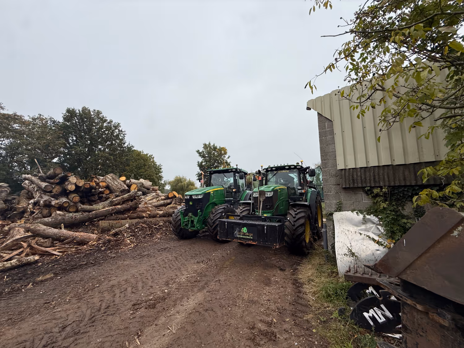 Twee groene tractoren staan geparkeerd op een onverhard pad naast een grote stapel gekapte boomstammen en een golfplaten schuur, onder een bewolkte hemel met bomen rondom.