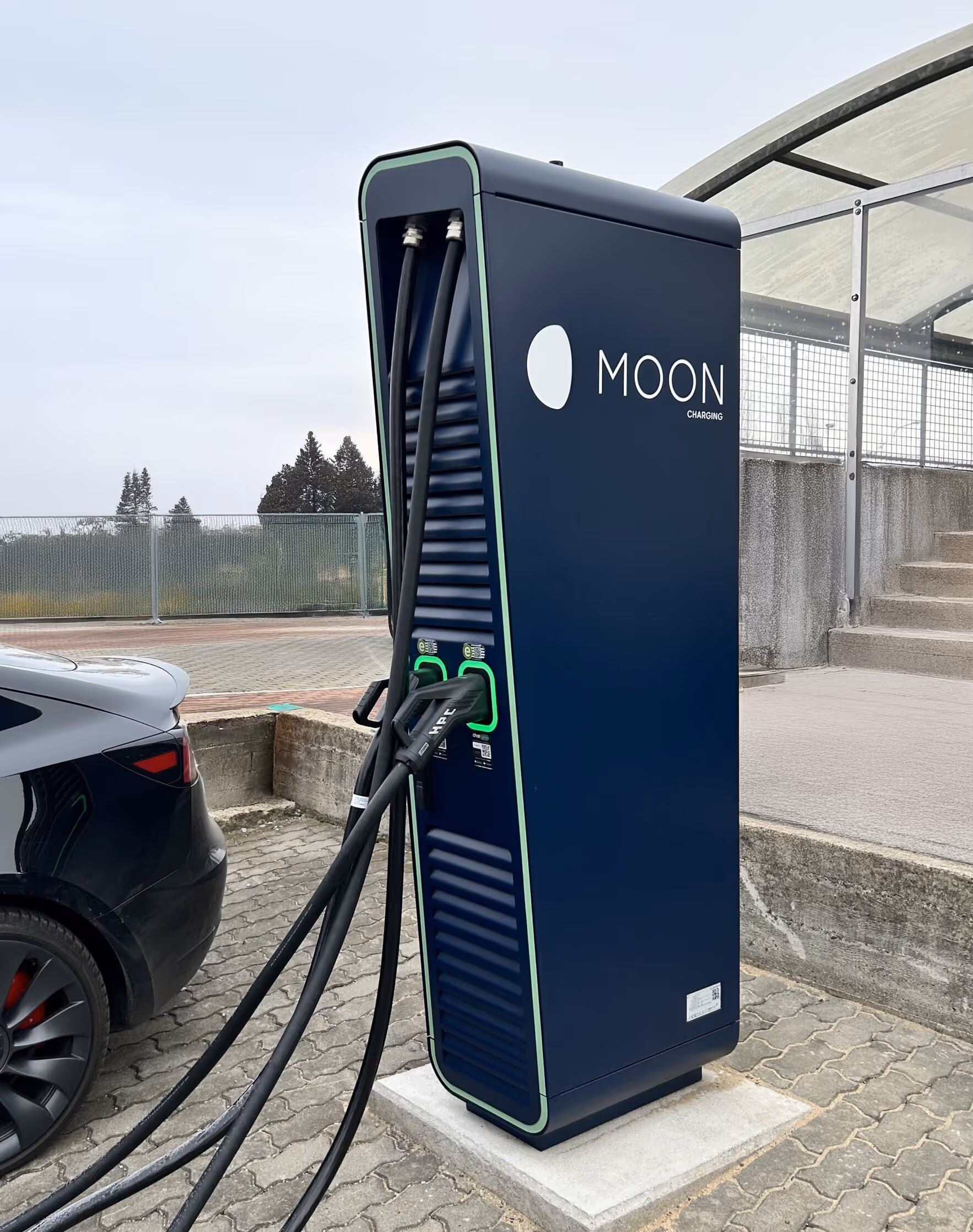 Černá elektrická nabíjecí stanice s nápisem MOON u auta Tesla na parkovišti.