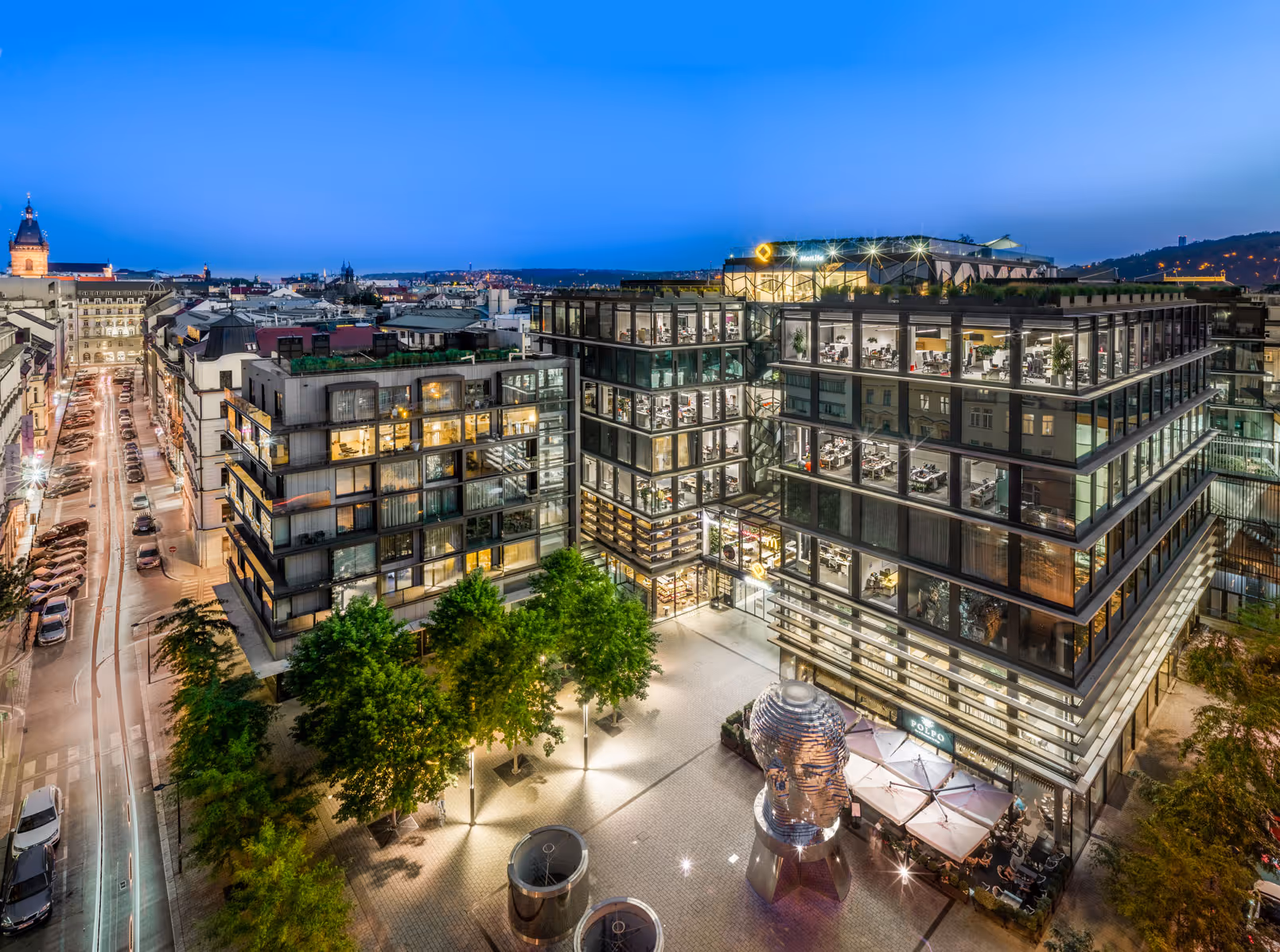 Noční městská scéna s moderní kancelářskou budovou s prosklenými stěnami, osvětleným náměstím a sochou hlavou z kovových vrstev.