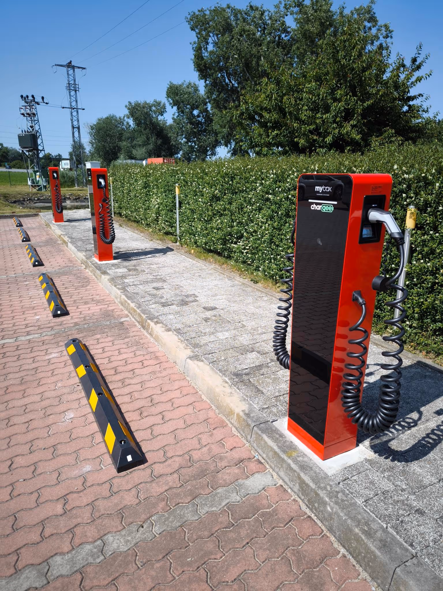 Tři červené nabíjecí stanice pro elektromobily podél chodníku vedle keře za slunečného dne.