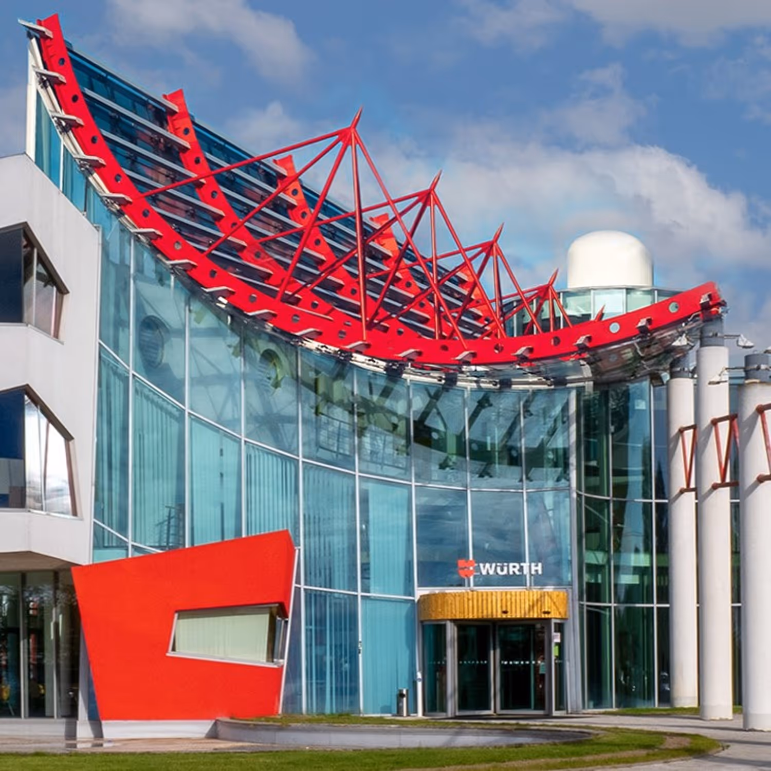 Moderní budova s velkými skleněnými stěnami, červenými kovovými prvky a vchodem se značkou Würth.