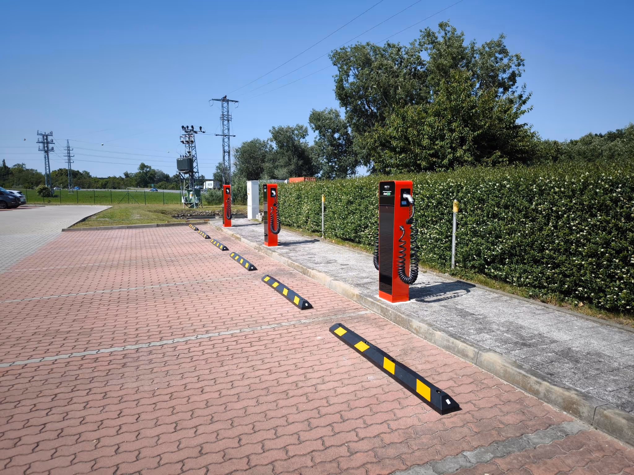 Parkoviště s červenými stojany pro nabíjení elektrických vozidel vedle zeleného živého plotu za slunečného dne.