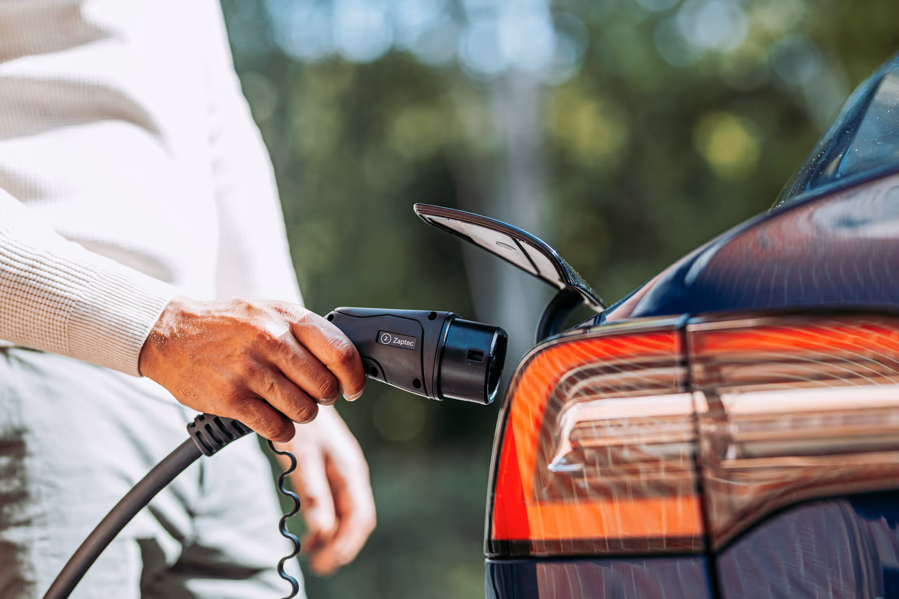 Ruka drží nabíjecí konektor připojený k elektromobilu modré barvy.