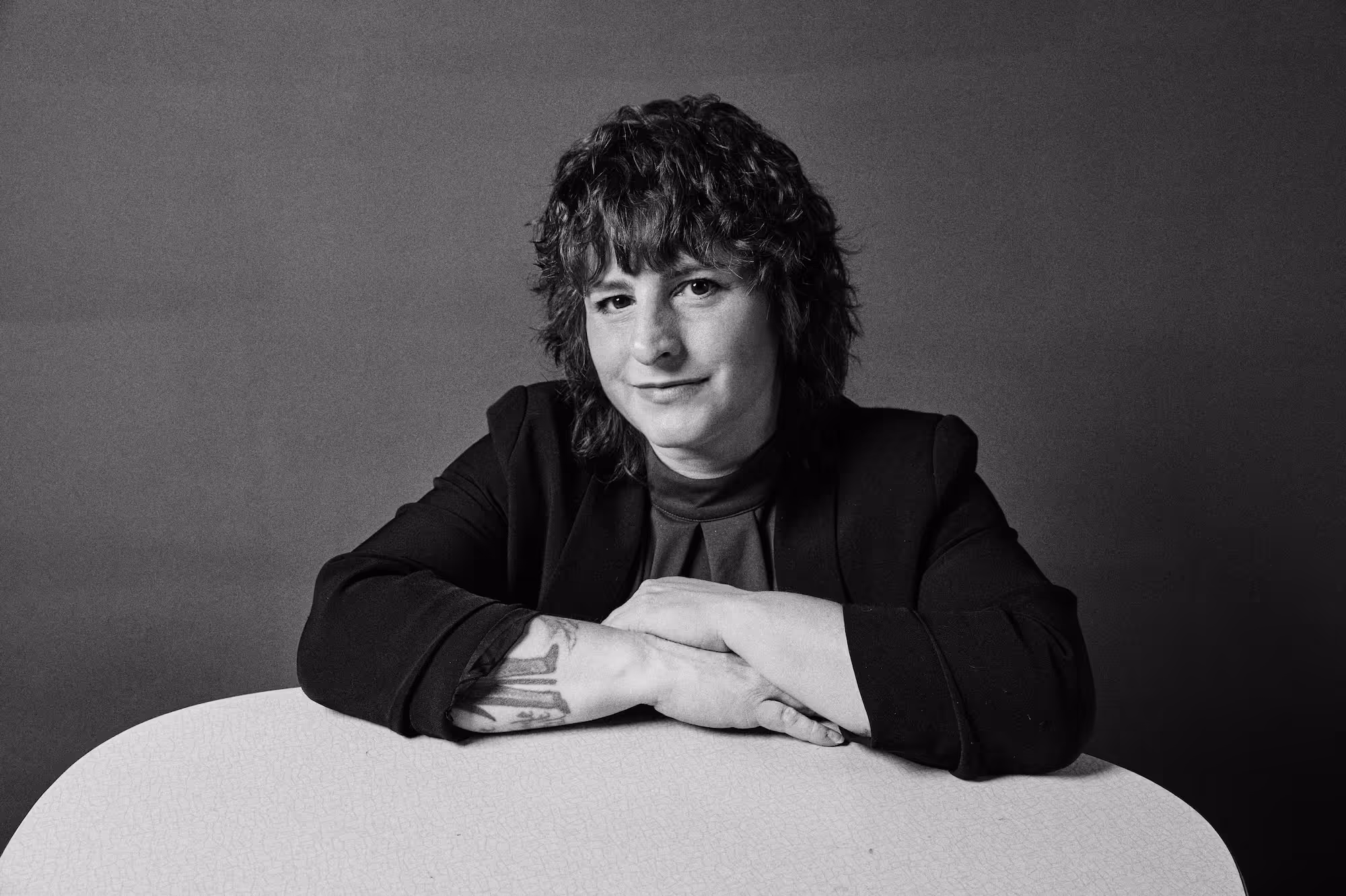 Schwarz-weiß-Porträt einer Person mit lockigem Haar, die lächelnd am Tisch sitzt, die Arme gekreuzt und ein Tattoo am Unterarm zeigt.