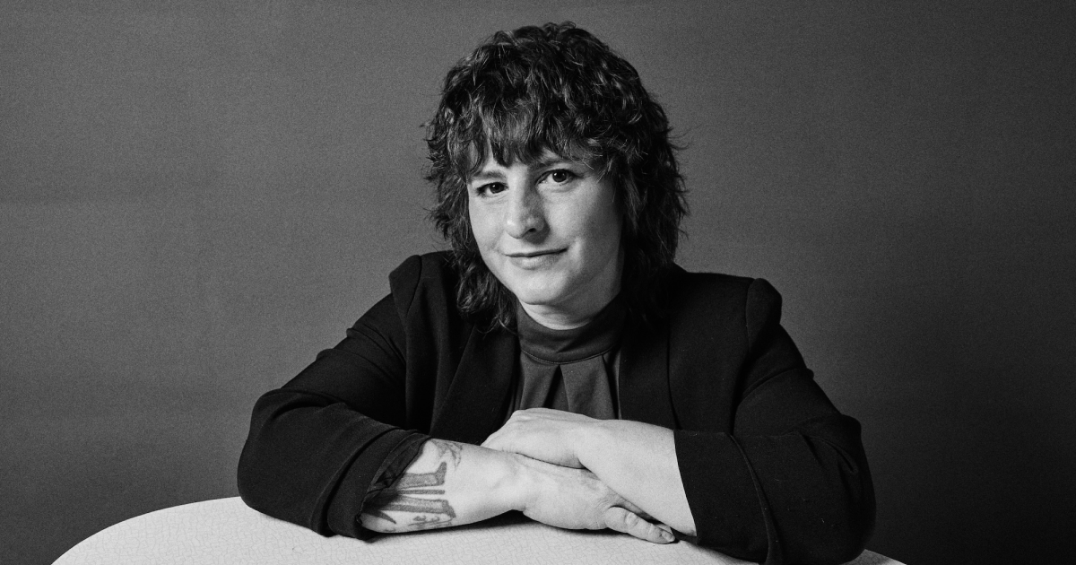 Schwarz-weiß-Porträt einer Frau mit gewelltem Haar, die an einem Tisch sitzt, mit verschränkten Armen und sichtbarem Tattoo am Unterarm.