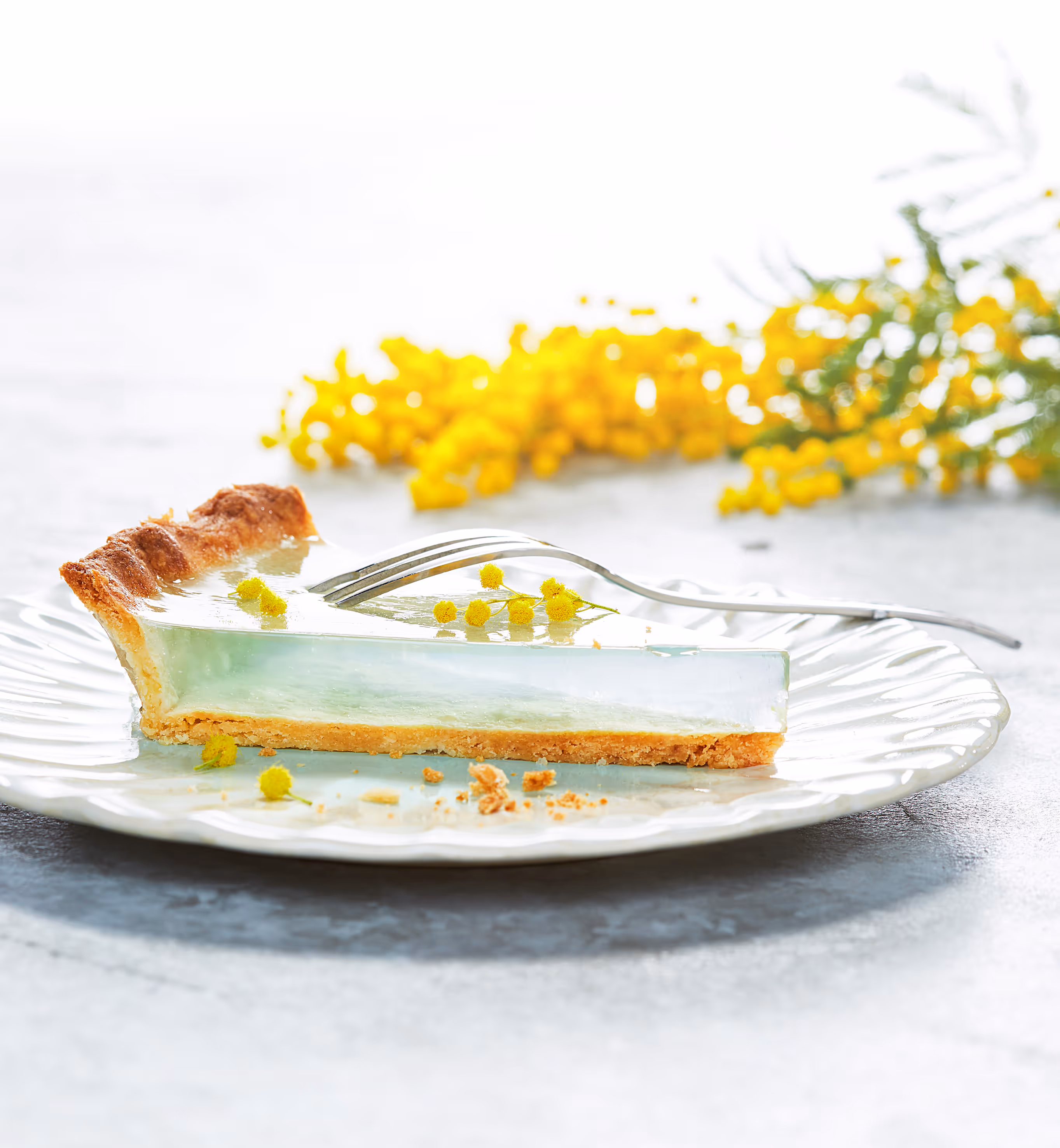 Ein Stück klarer Geleekuchen mit knusprigem Mürbeteigboden auf einem weißen Teller, dekoriert mit kleinen gelben Blüten und einer silbernen Gabel.