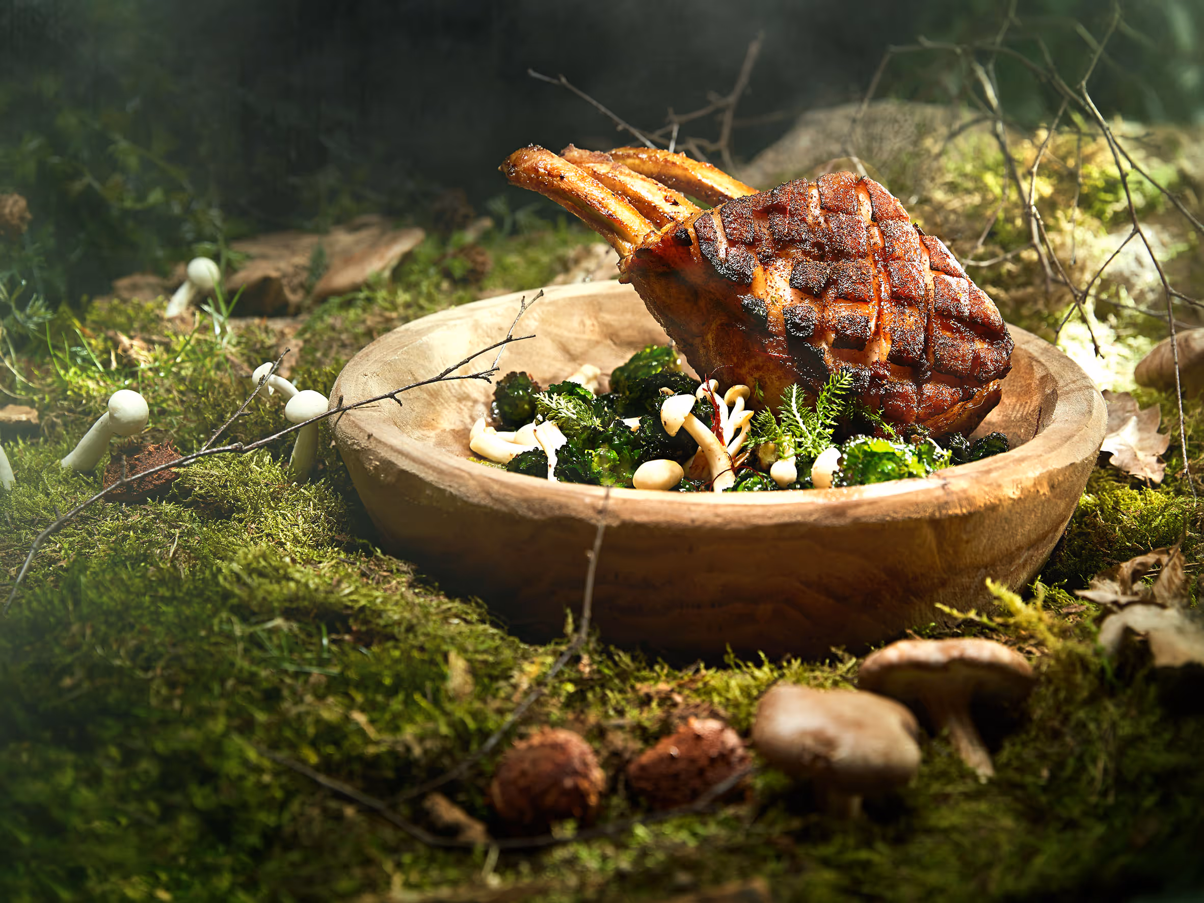 Gegrilltes Fleisch mit knuspriger Kruste in einer Holzschale, umgeben von grünem Gemüse und Pilzen auf moosigem Waldboden.