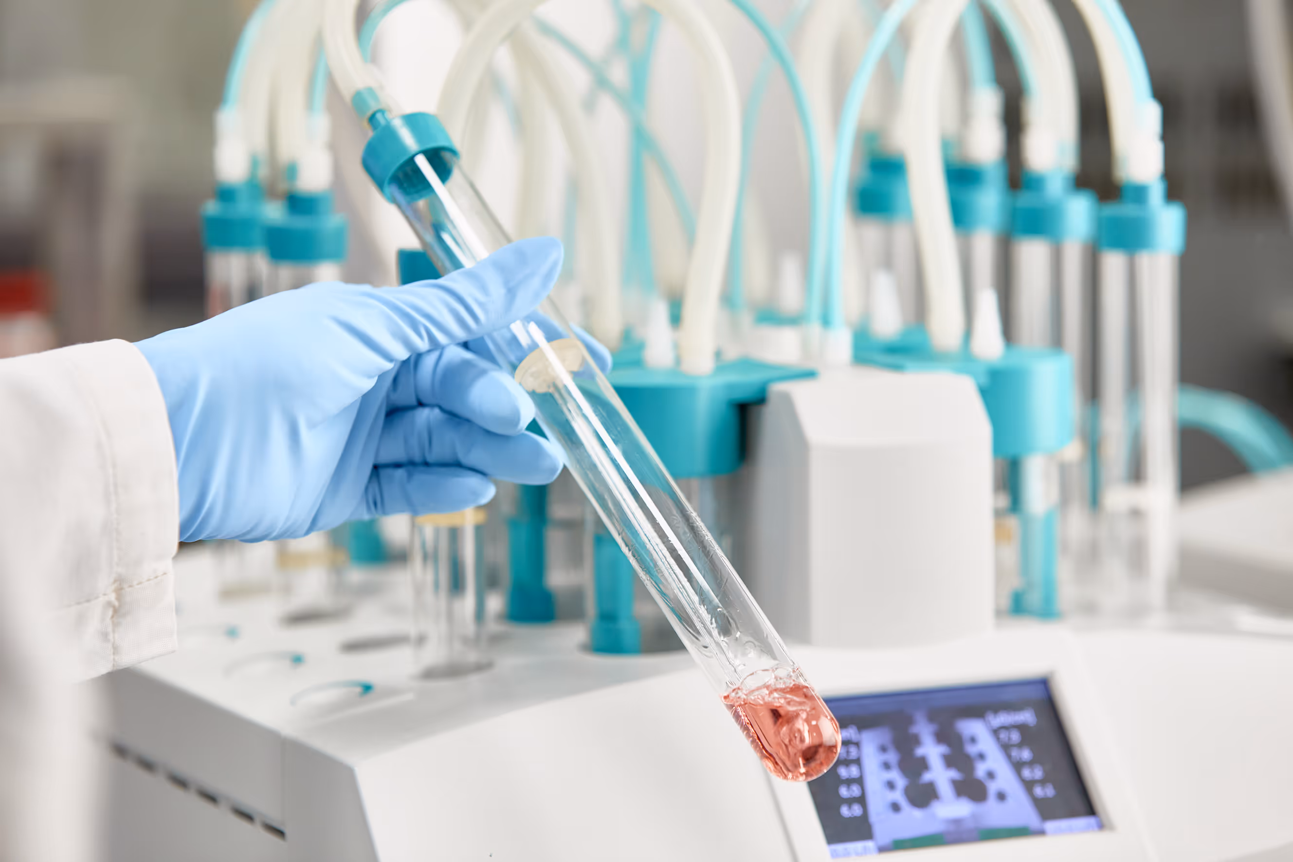 Unternehmen Fotografie in einem Labor. Hand mit blauen Handschuhen hält ein Reagenzglas mit rosafarbener Flüssigkeit vor einem Laborgerät mit Schläuchen und digitalem Display.