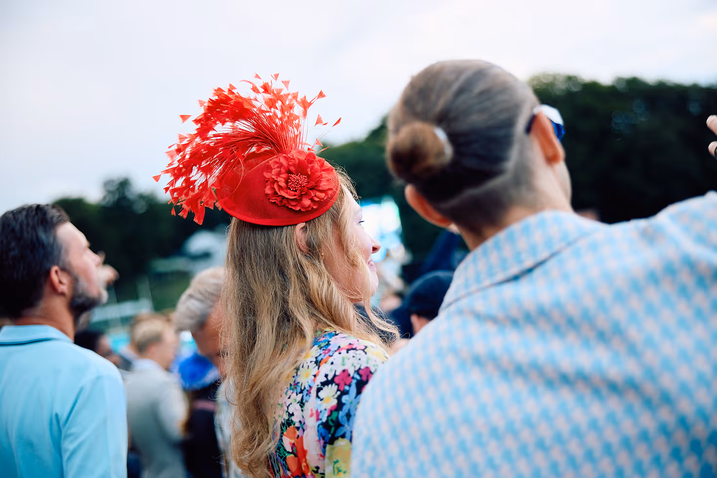 Woman in a colorful floral dress wearing a decorative red fascinator at an outdoor event.