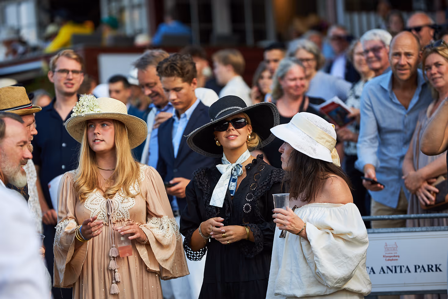 Three women wearing elegant hats and dresses at an outdoor event with a crowd behind them.