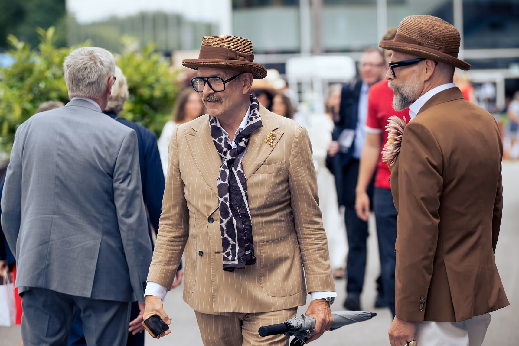 Two men in vintage suits and hats, one holding an umbrella and phone, on a city street with crowds.