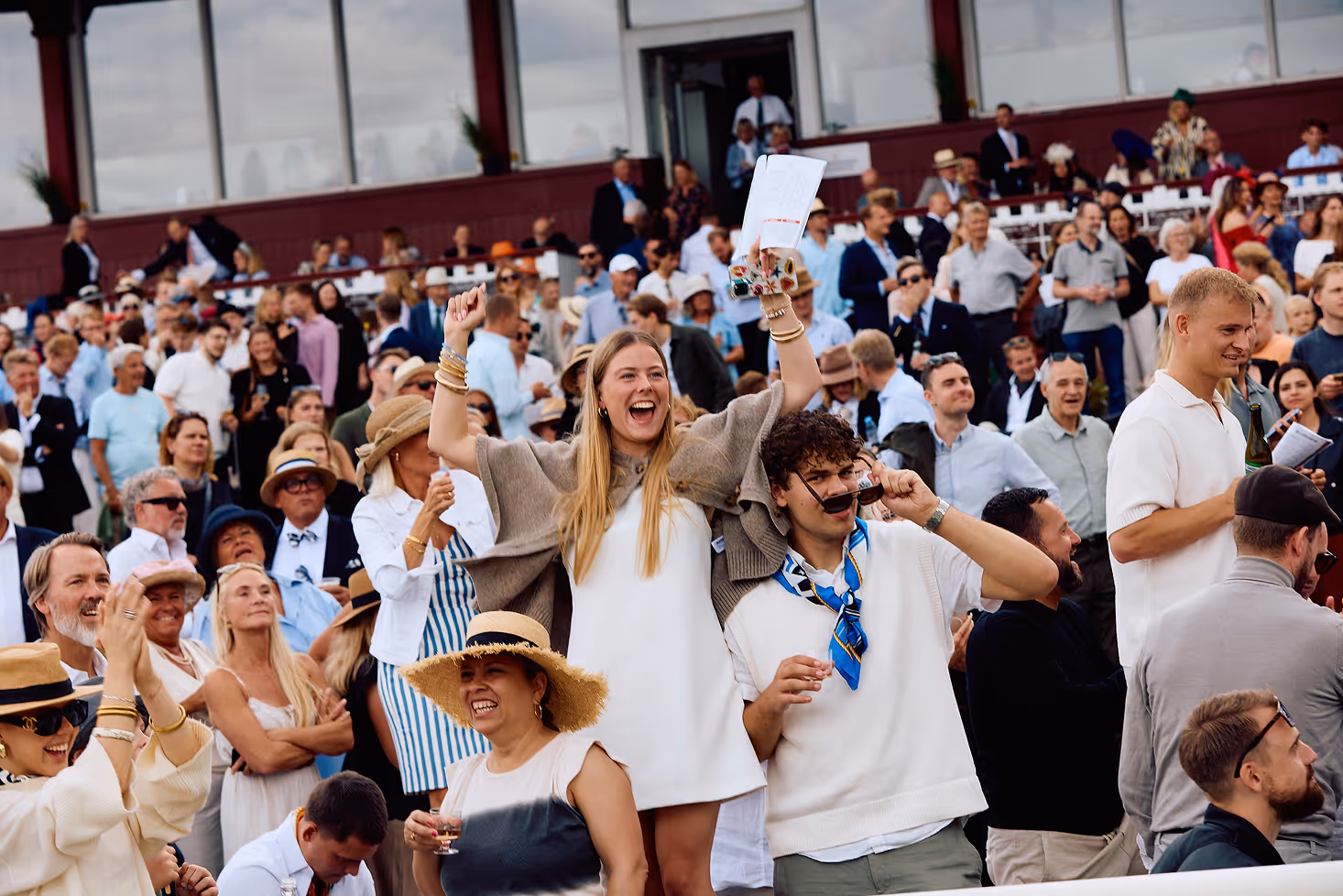 Crowd at event cheering, with a woman raising arms excitedly and a man adjusting sunglasses nearby.