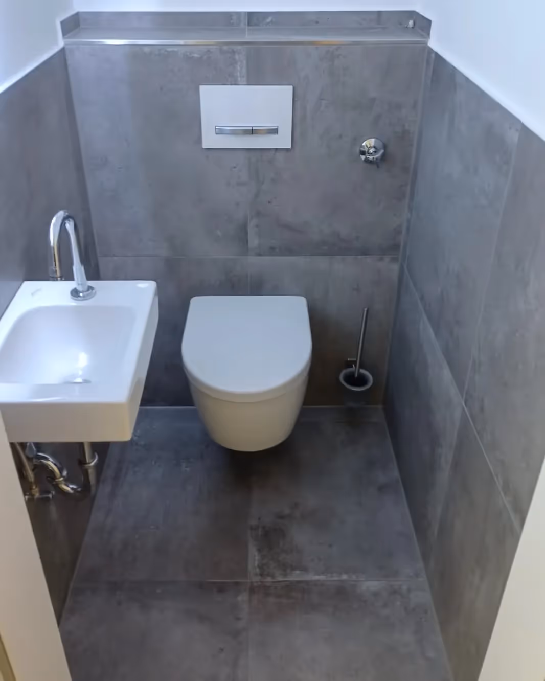 Kleines modernes Badezimmer mit wandhängendem WC, kleinem weißen Waschbecken und grauen Wandfliesen.