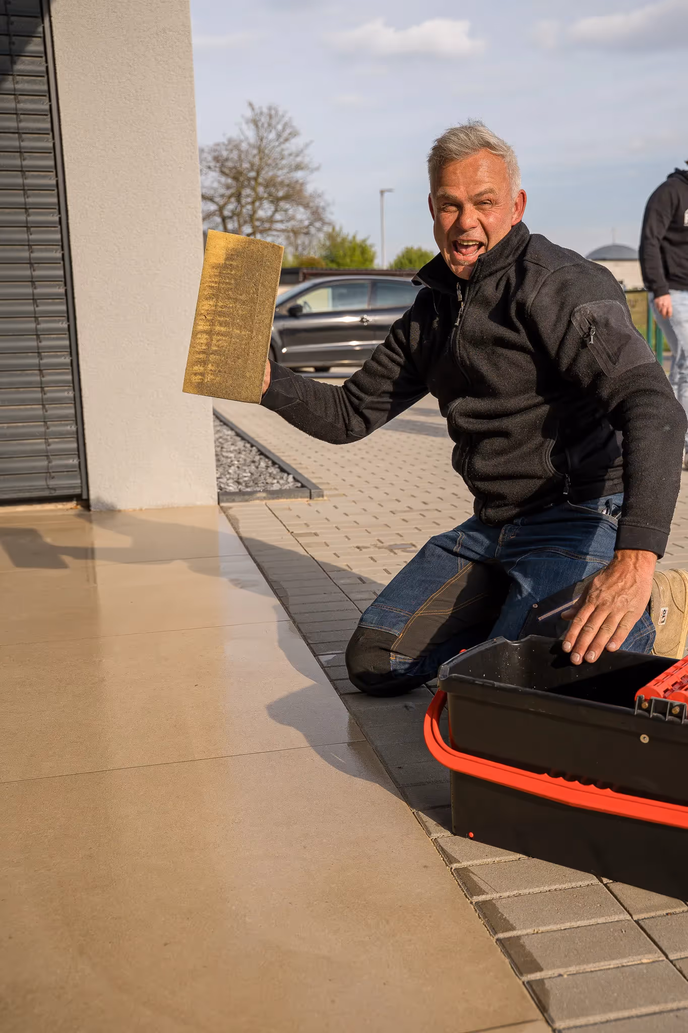 Mann kniet draußen auf dem Boden und hält einen Schwamm, daneben eine Werkzeugbox mit roten Griffen.