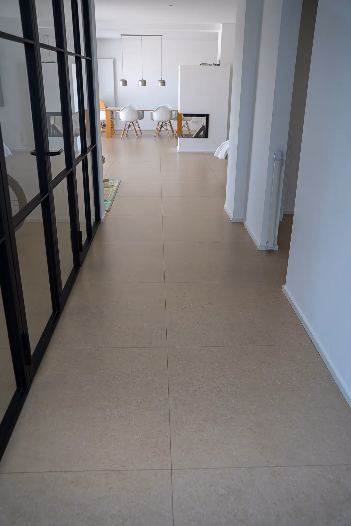 Flur mit beigen Bodenfliesen, schwarzer Glastür links, weißem Kamin und weißem Esstisch mit vier Stühlen im Hintergrund.