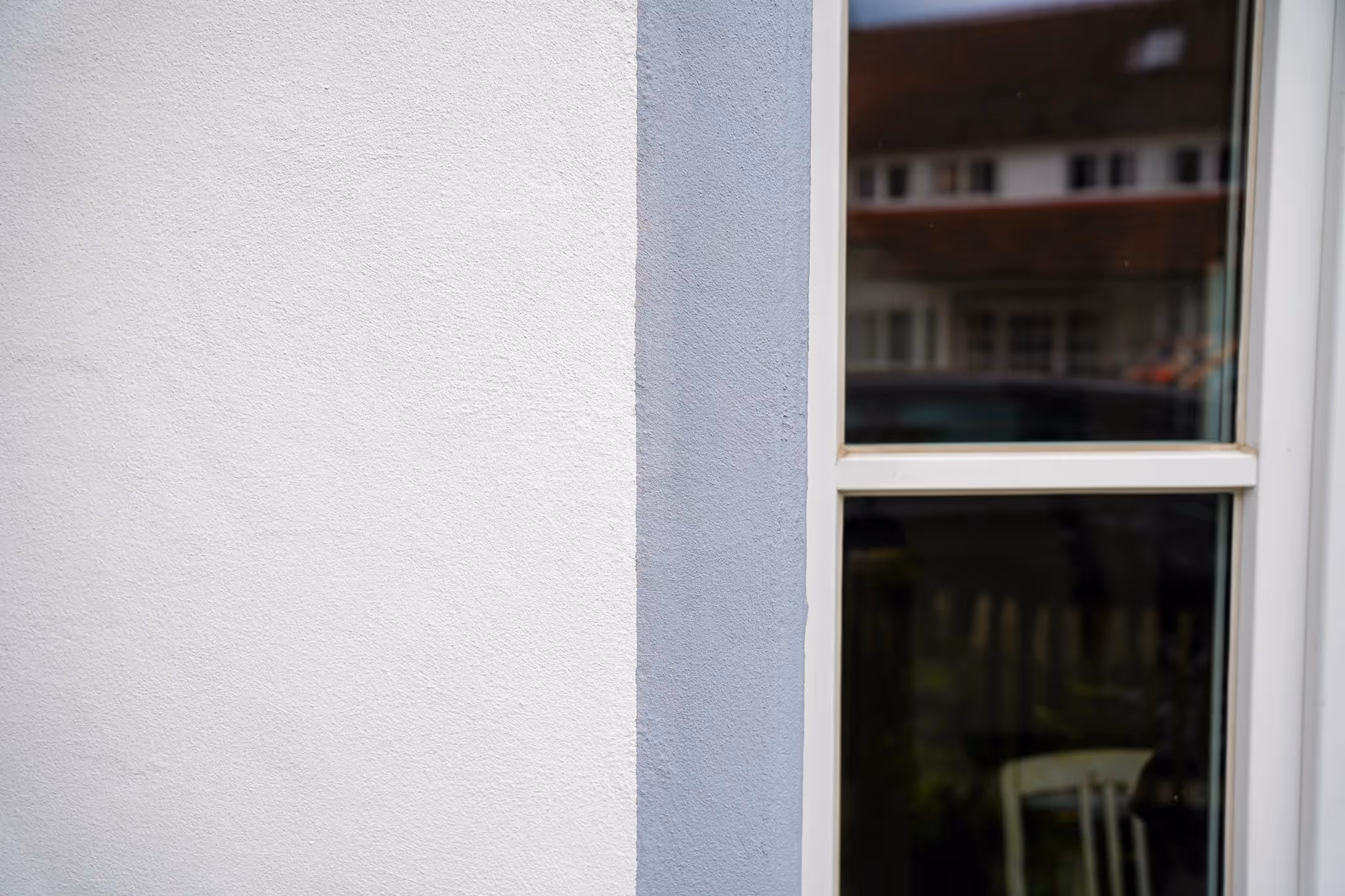 Detailansicht eines weiß-grau gestrichenen Gebäudewandabschnitts neben einem Fenster mit reflektierendem Glas.