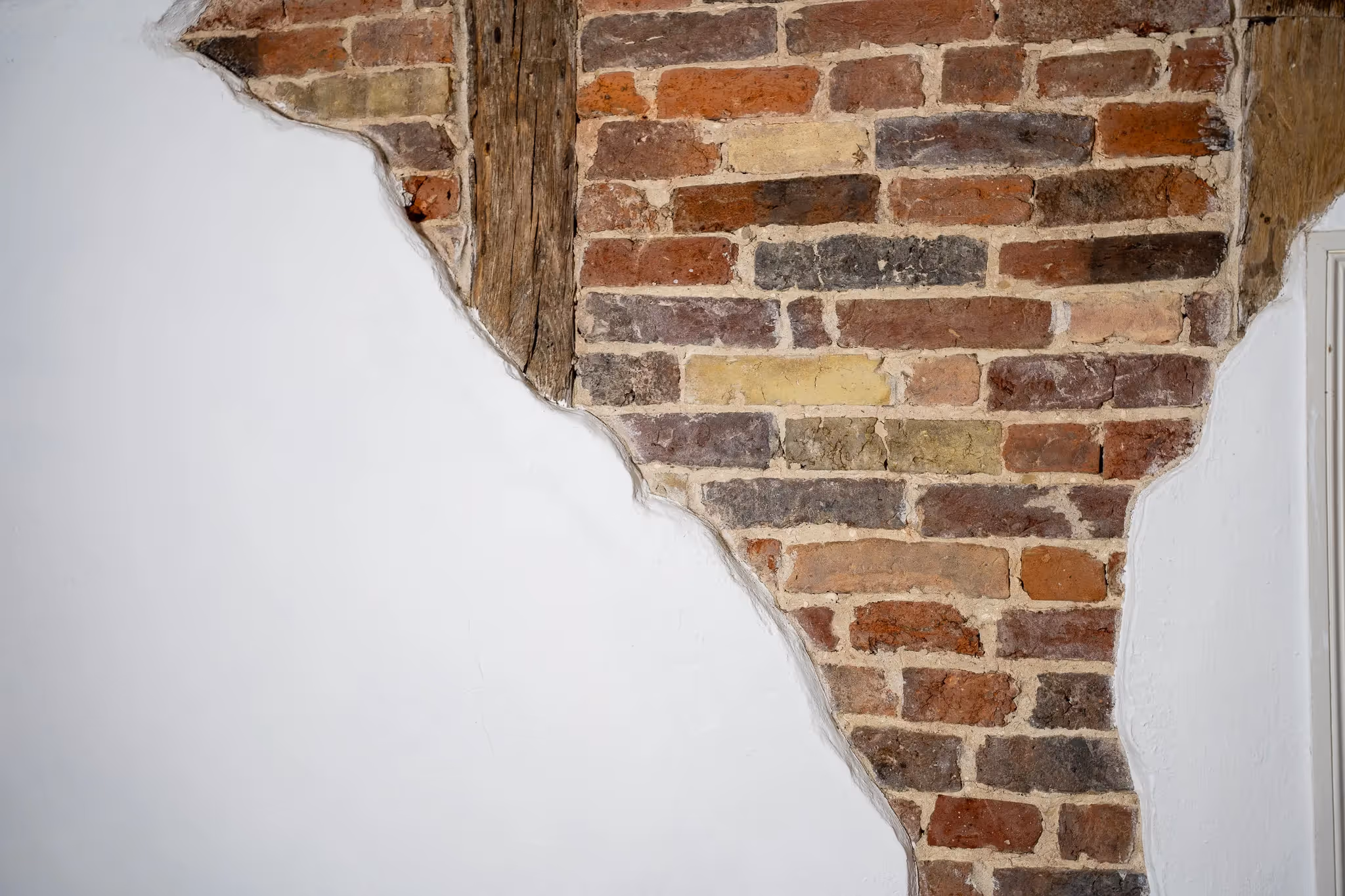 Teilweise freigelegte Backsteinwand mit einem vertikalen Holzträger, umgeben von weiß verputzter Wand.