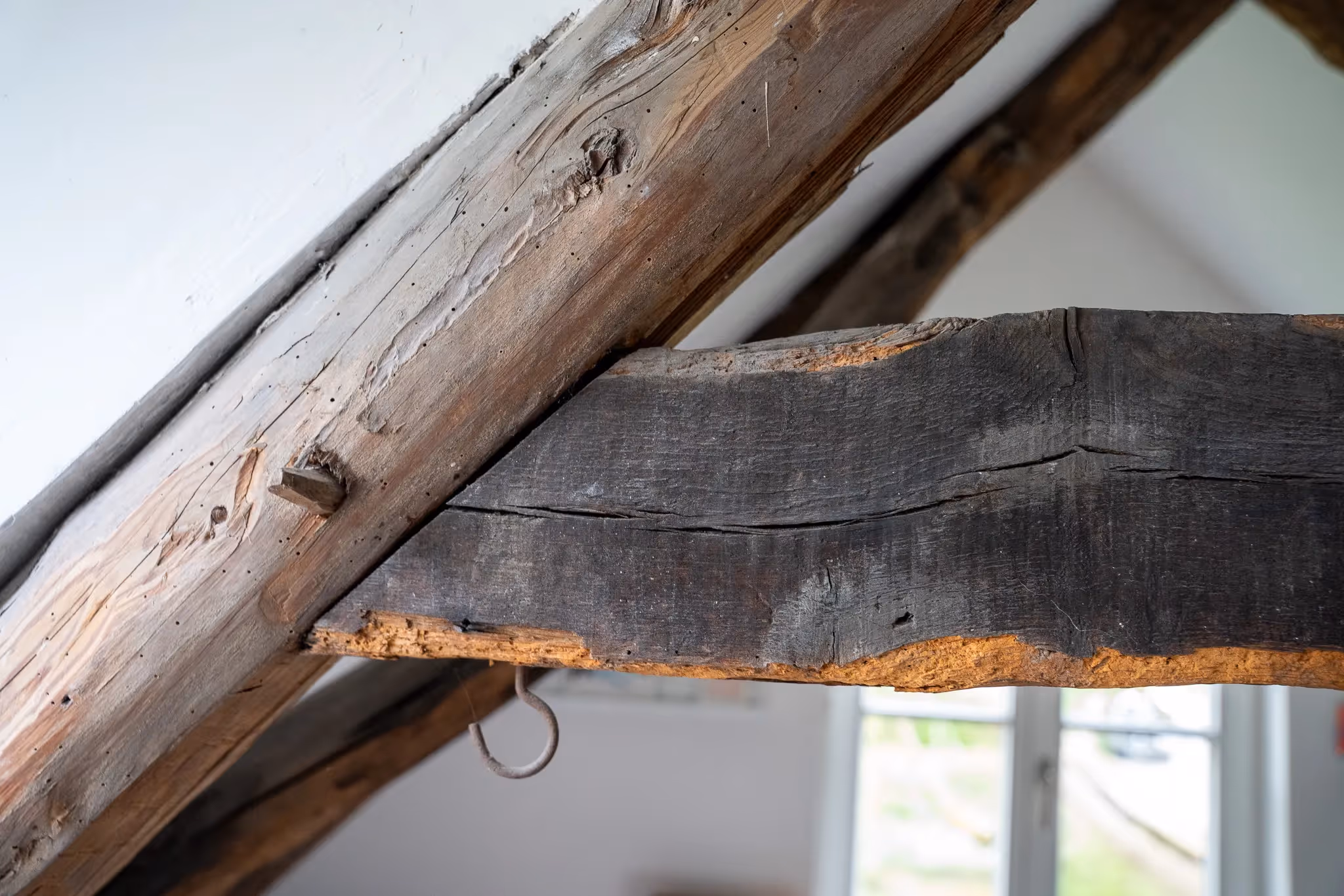 Nahaufnahme von verwittertem Holzbalken und Dachbalken in einem Raum mit Fenster im Hintergrund.