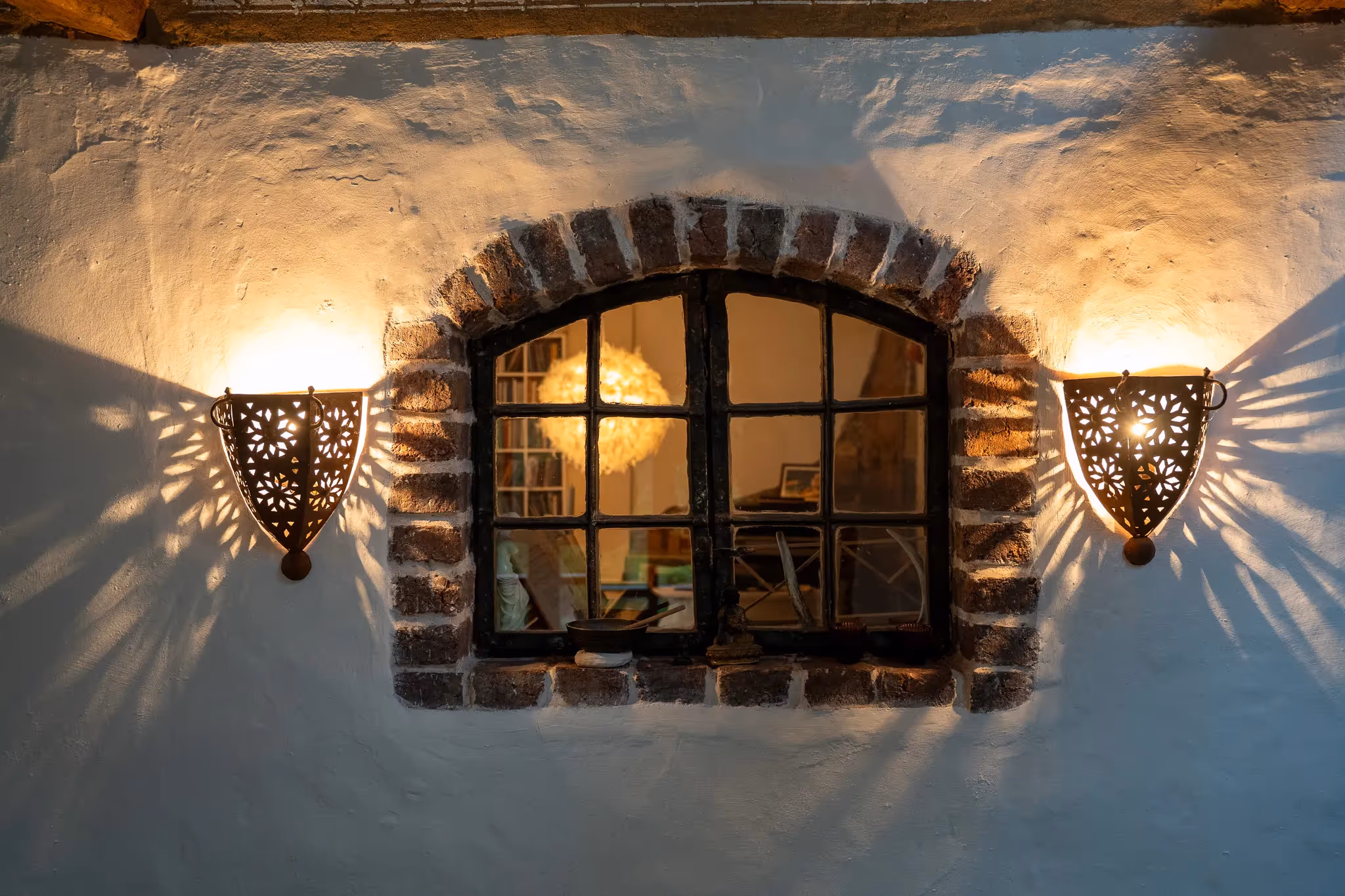 Beleuchtetes Fenster mit gemauertem Rundbogen und zwei dekorativen Wandlampen, die Schatten an die Wand werfen.