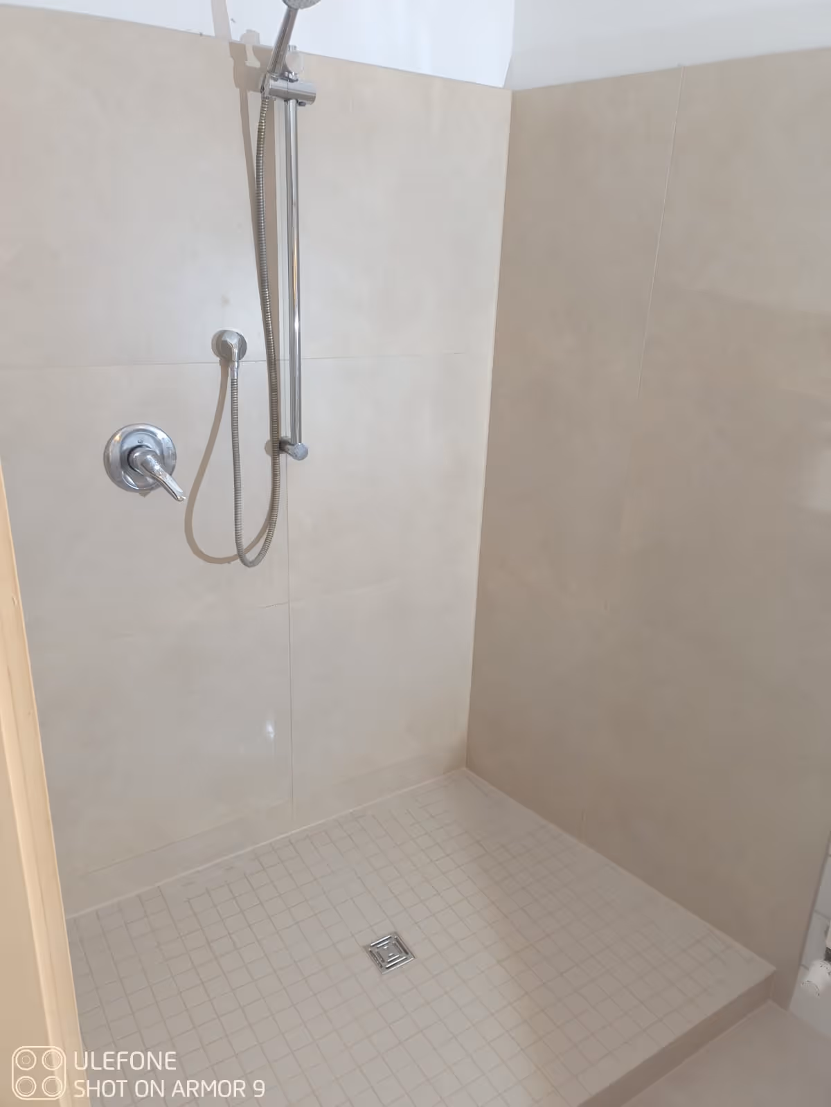 Minimalistische Dusche mit beigen Fliesen und verstellbarem Duschkopf an einer Stange.