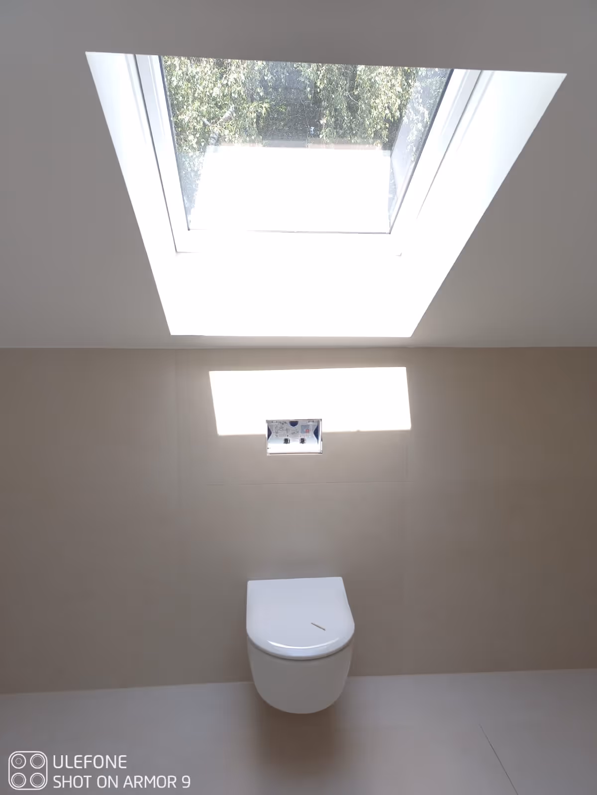 Minimalistisches Badezimmer mit einer an der Wand montierten Toilette unter einem großen Dachfenster, durch das Tageslicht fällt.