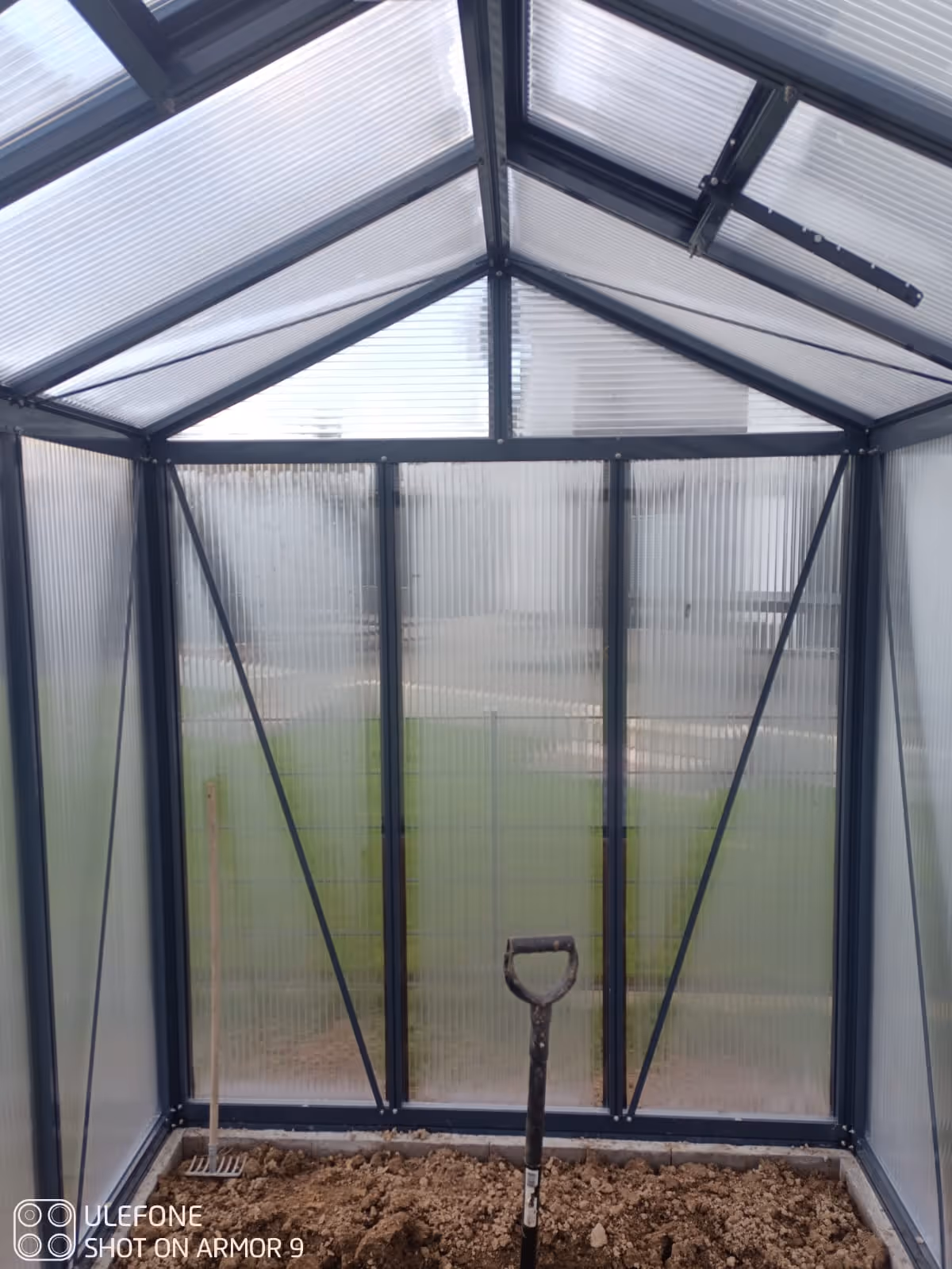 Innenansicht eines leeren Kleingewächshauses mit Boden aus Erde und zwei Gartenwerkzeugen, einer Hacke und einer Schaufel.