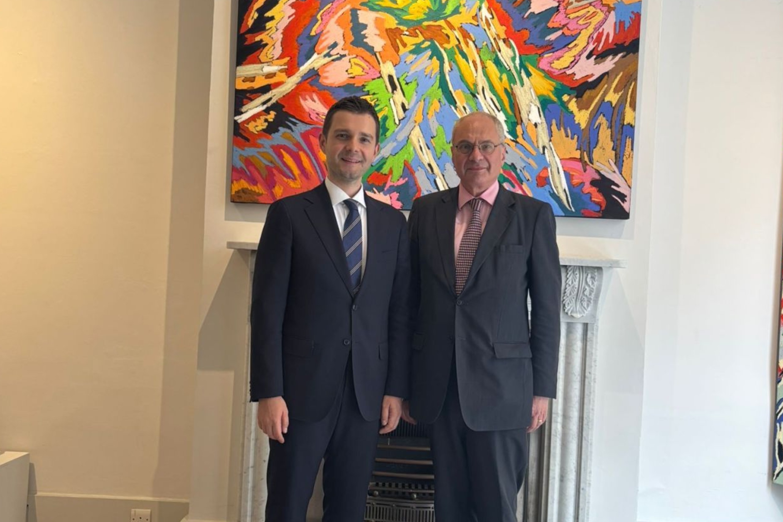 Two men in dark suits standing side by side in front of a colorful abstract painting and white fireplace.