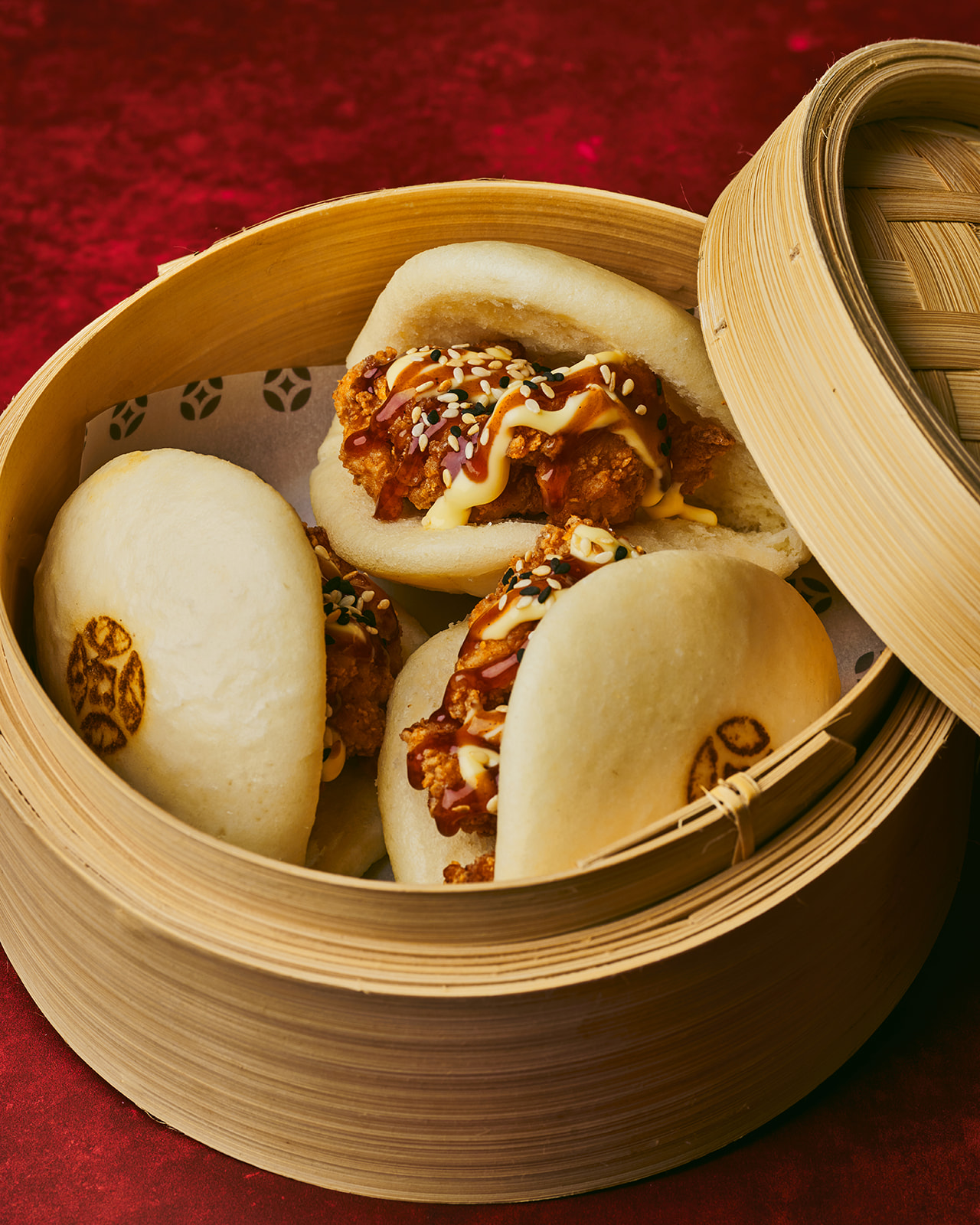 Steamed bao buns filled with crispy fried chicken, drizzled with sauces and sprinkled with sesame seeds in a bamboo steamer basket.