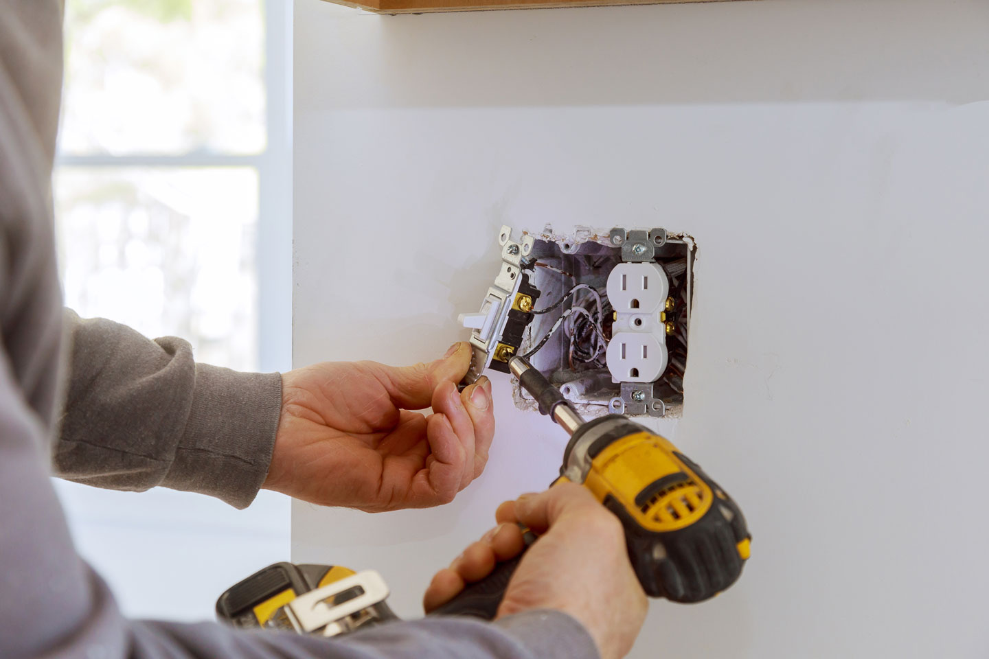 Person installing a white electrical light switch next to a dual electrical outlet, using a cordless drill.