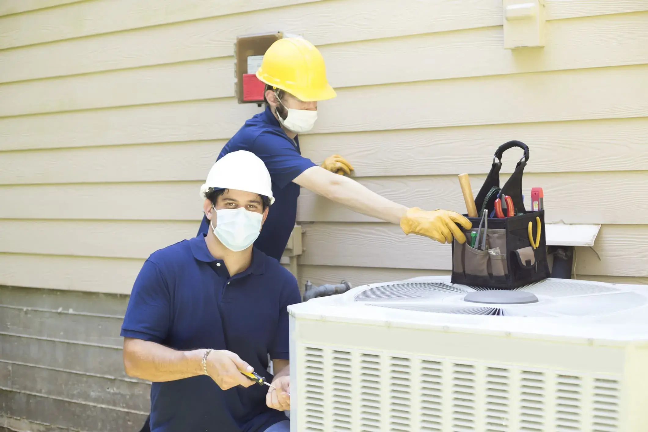 Two workers in protective masks and yellow hard hat repairing outdoor air conditioning unit