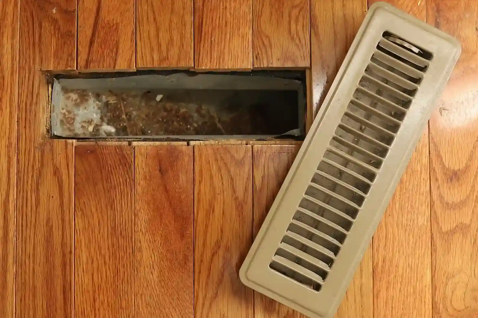 Open rectangular floor vent with beige cover beside it, showing debris inside on wooden flooring