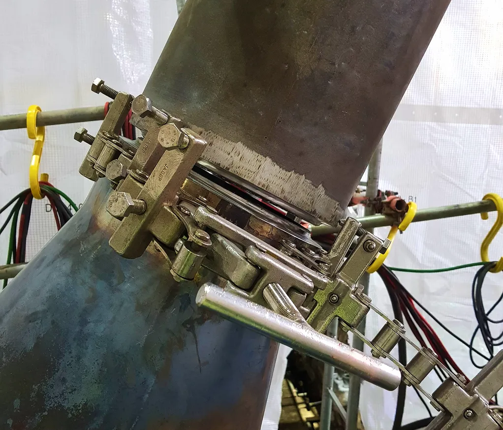 Nahaufnahme eines großen Metallrohranschlusses, an dem eine präzise Justier- und Spannvorrichtung für die Schweißvorbereitung befestigt ist; das Rohr ist im Nahtbereich metallisch blank geschliffen und von Gerüst- und Kabelinstallationen umgeben.