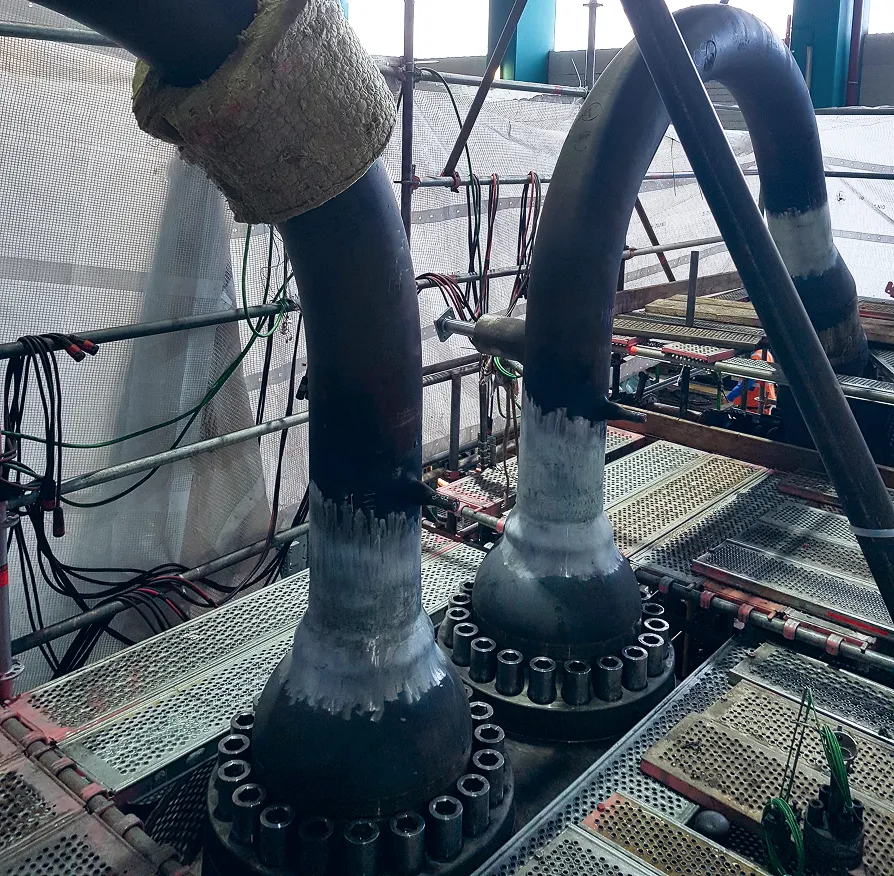 Industrielle Anlage mit zwei großen gebogenen Rohren, deren Verbindungsbereiche für das Schweißen vorbereitet sind: blank geschliffene Zonen, umgeben von Gerüsten und technischem Equipment.“