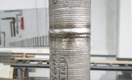 Nahaufnahme eines Metallrohrs mit einer umlaufenden Schweißnaht in der Mitte; geriffelte Oberfläche, ungleichmäßige Schweißraupe, im Hintergrund eine industrielle Werkstatt mit Metallkonstruktionen und Maschinen.
