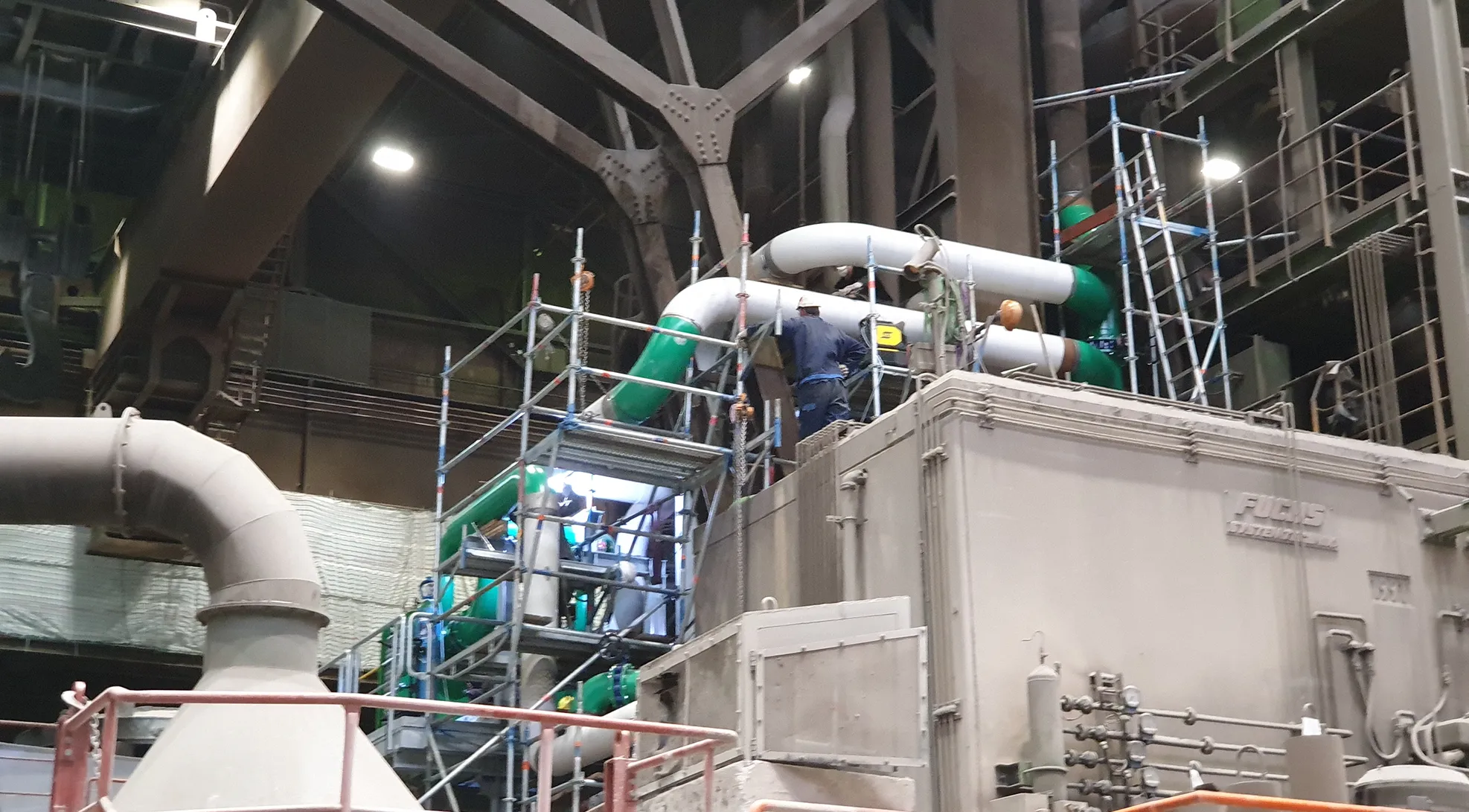 Montagearbeiten an großen Industrie‑Rohrleitungen auf Gerüst in einer Anlagehalle.