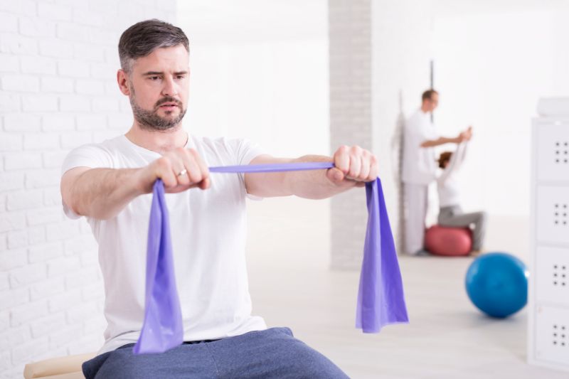 A person is doing physical therapy with a purple resistance band, with other people in the background also engaged in therapy exercises.