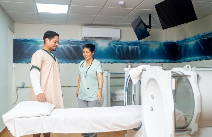 A healthcare professional in scrubs and a patient in a tan gown stand near a medical bed and a hyperbaric oxygen therapy chamber with ocean wave mural on the wall.