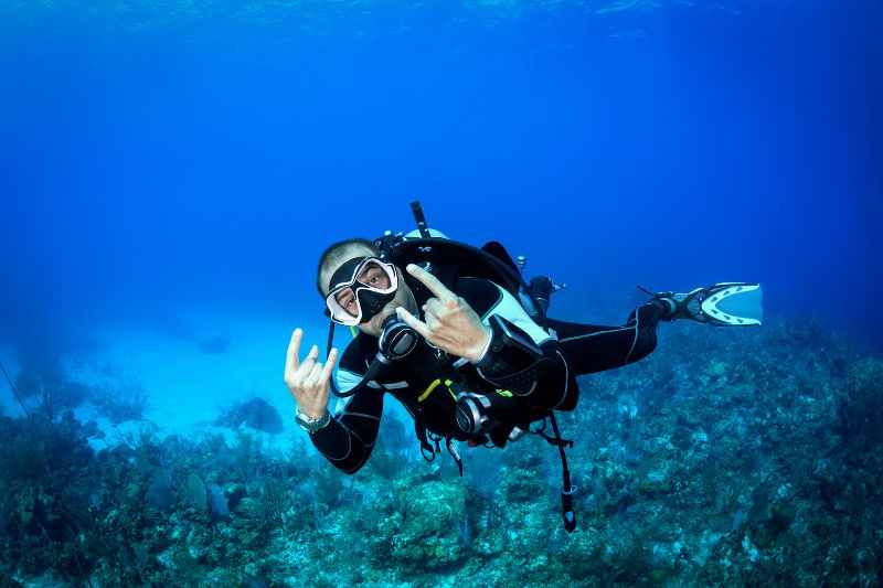 A scuba diver in full gear gives a rock-and-roll hand gesture underwater near a coral reef.
