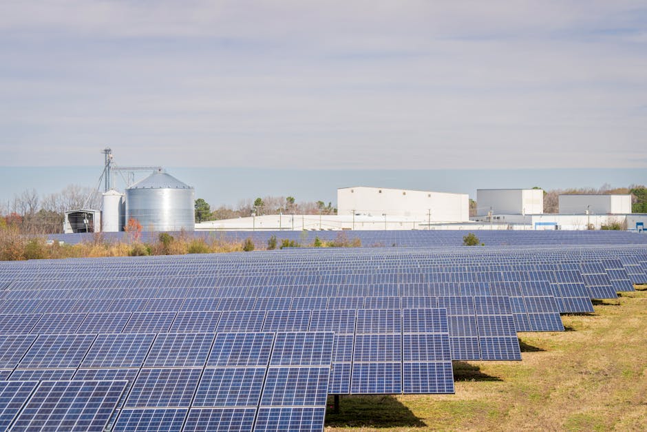 A large solar panel field with warehouses and silos in the background under a clear sky.