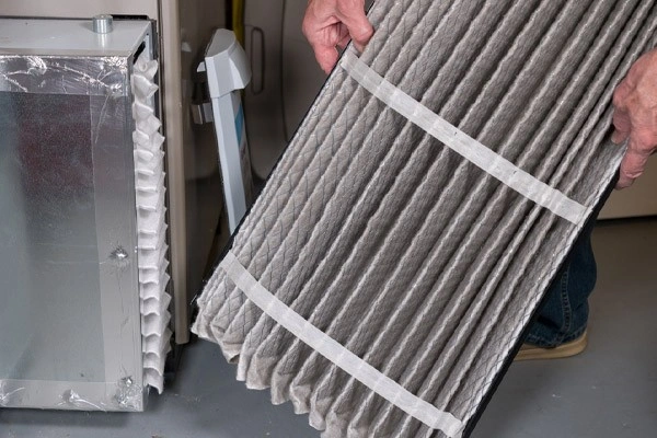 Hands holding a dirty, pleated air filter next to an HVAC unit