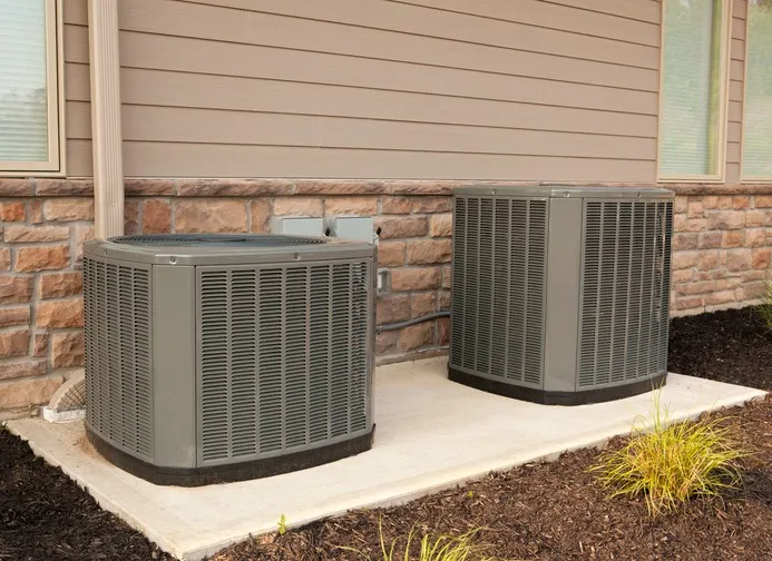 Two gray air conditioning units outside a brick and siding house