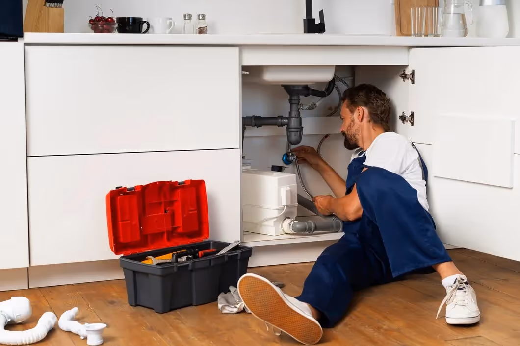 Plumber repairing sink pipes under kitchen cabinet with toolbox nearby