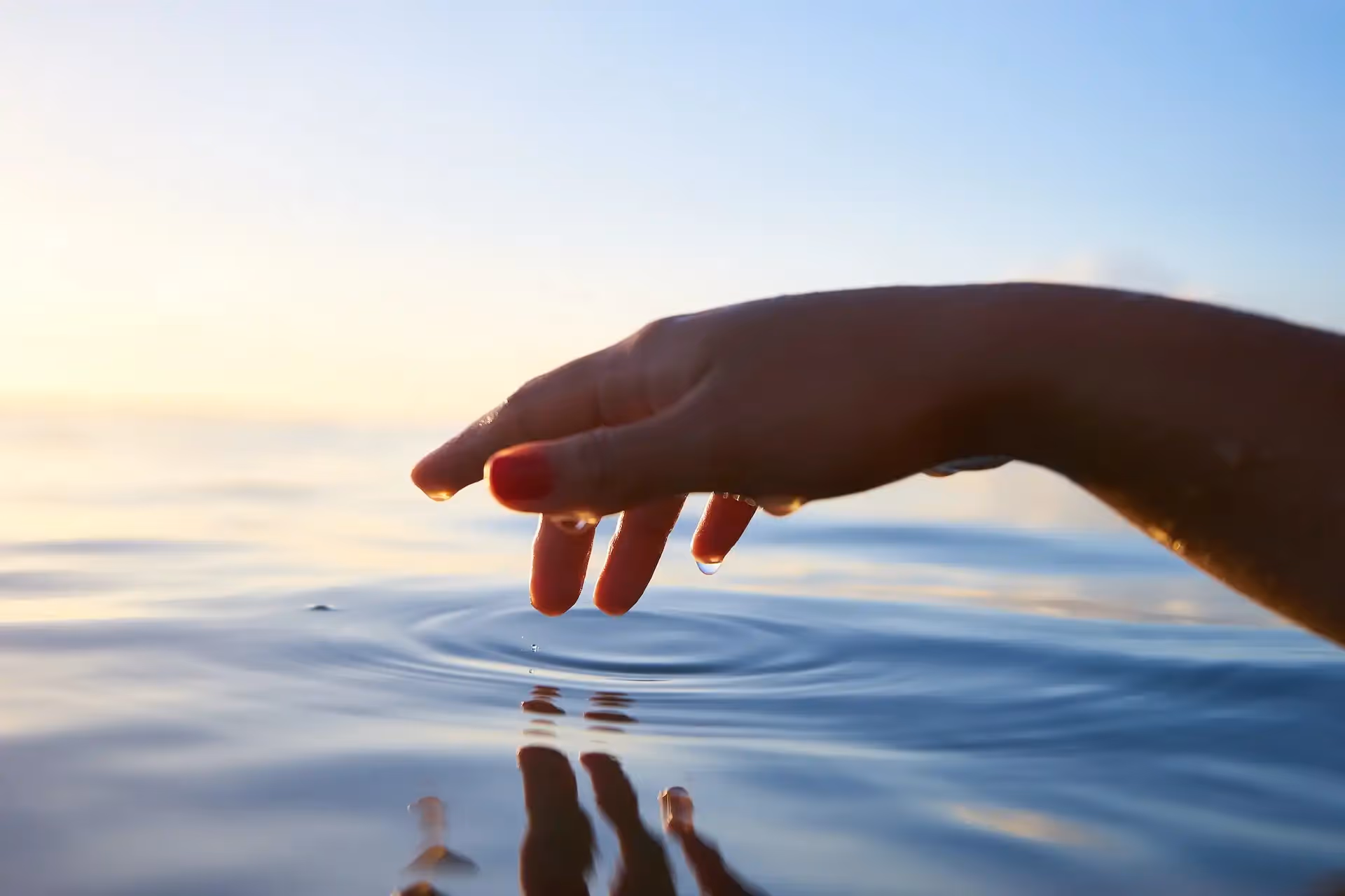 Hand touching water surface, creating ripples at sunset