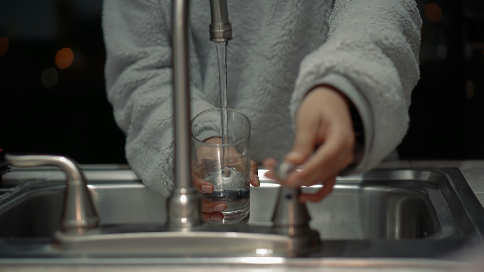 Person in cozy sweater filling glass with water from kitchen faucet